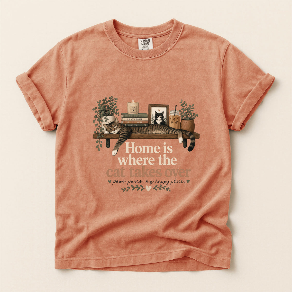 A peach-colored short-sleeve t-shirt with a graphic of a cat lounging on a shelf with books and plants, accompanied by the text 'Home is where the cat takes over' and 'paws, purrs, my happy place' on a light beige background.